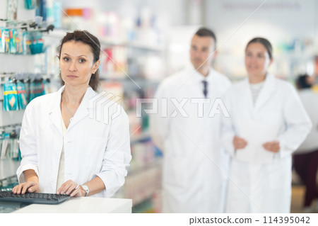Middle-aged female pharmacist standing at the desk using keyboard in chemist's shop Middle-aged female pharmacist standing at the desk using keyboard in chemist's shop 114395042