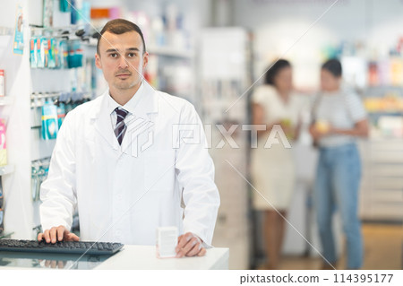 Young male pharmacist standing at the desk using keyboard in chemist's shop Young male pharmacist standing at the desk using keyboard in chemist's shop 114395177