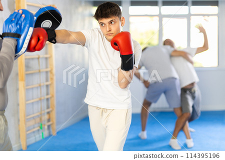 Coach holding mitts during self defence training in gym with young guy mastering base punches in boxing gloves in gym Coach holding mitts during self defence training in gym with young guy mastering base punches in boxing gloves in gym 114395196