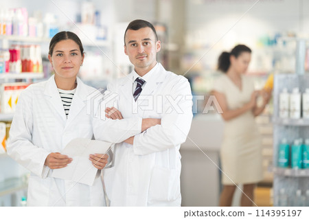 Positive man and girl pharmacists are standing in pharmacys sales hall, pharmacy staff are amicably preparing to meet chemist shop visitors Positive man and girl pharmacists are standing in pharmacys sales hall, pharmacy staff are amicably preparing to meet chemist shop visitors 114395197