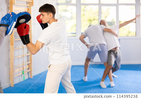 Coach holding mitts during self defence training in gym with young guy mastering base punches in boxing gloves in gym Coach holding mitts during self defence training in gym with young guy mastering base punches in boxing gloves in gym 114395238