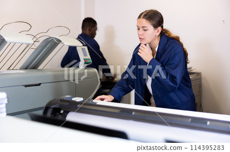 Focused young woman in uniform setting up the plotter before starting work in the printing house 114395283
