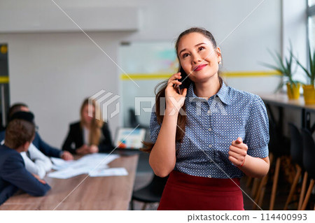 Asian businesswoman in smart casual attire engages in phone communication, assured by roaming connectivity in bustling corporate office environment with multiethnic colleagues working in blur at back. 114400993