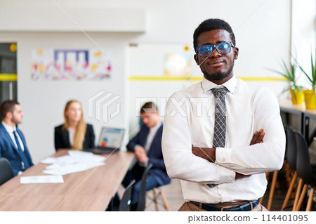 Confident African businessman stands in modern office, arms crossed. Teamwork in background, inclusive and diverse work environment. Pro attire, young leader, business strategy session. Confident African businessman stands in modern office, arms crossed. Teamwork in background, inclusive and diverse work environment. Pro attire, young leader, business strategy session. 114401095