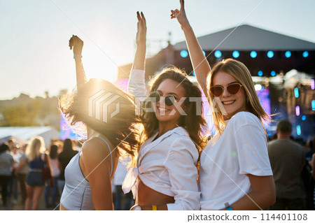 Group of joyful young women dancing at sunlit beach music fest. Friends enjoy live concert, summer vibes, hands up in air. Trendy girls fun weekend on sandy shore with festival stage in background. 114401108