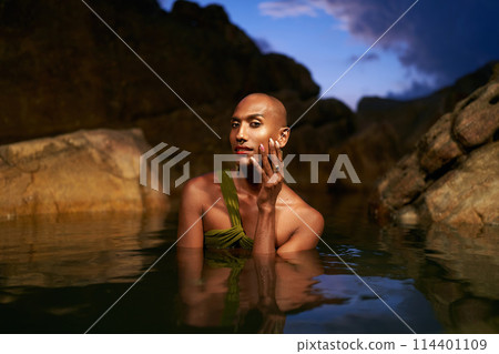 Gay black lgbtq man dives out of water inside picturesque natural rocky pond at night. Homosexual biethnic person poses waist deep in still water among stones on exotic island. Pride month LGBTQIA. Gay black lgbtq man dives out of water inside picturesque natural rocky pond at night. Homosexual biethnic person poses waist deep in still water among stones on exotic island. Pride month LGBTQIA. 114401109