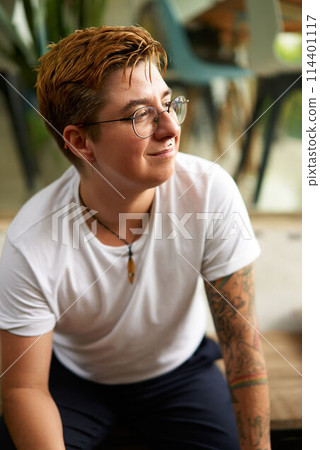 Positive transgender individual in casual setting, smiling confidently. Short hair, tattoo embodies inclusion, empowerment in modern society. Gender identity, workplace diversity. 114401117