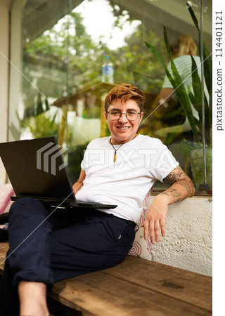 Transgender pro smiles while working on laptop in casual office space. Relaxed attire, inclusive work environment evident. Barefoot, shows comfort, tech engagement and confidence. 114401121