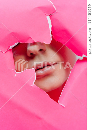View of beautiful young woman with plumped lips through hole in pink paper. View of beautiful young woman with plumped lips through hole in pink paper. 114401959