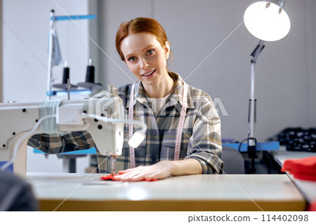redhead seamstress in factory sewing using industrial sewing machine, very accurate redhead seamstress in factory sewing using industrial sewing machine, very accurate 114402098