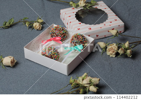 Cake Pops in chocolate with colored sprinkles. Decorated with a ribbon bow. They lie in a gift box, in the lid of which there is a transparent window. Nearby are dried roses. On a gray background. Cake Pops in chocolate with colored sprinkles. Decorated with a ribbon bow. They lie in a gift box, in the lid of which there is a transparent window. Nearby are dried roses. On a gray background. 114402212
