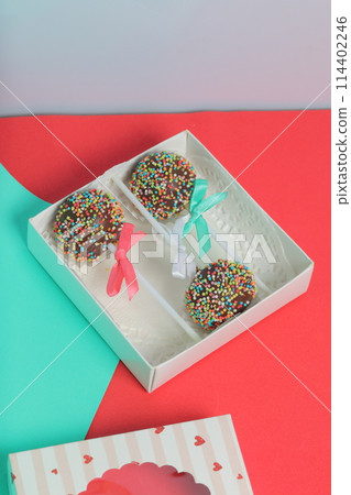 Cake Pops in chocolate with colored sprinkles. Decorated with a ribbon bow. They lie in a gift box, in the lid of which there is a transparent window. On a red and mint background. Cake Pops in chocolate with colored sprinkles. Decorated with a ribbon bow. They lie in a gift box, in the lid of which there is a transparent window. On a red and mint background. 114402246