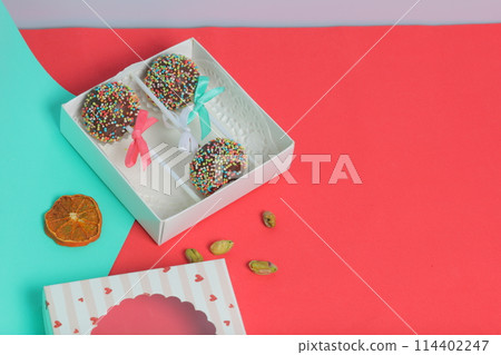 Cake Pops in chocolate with colored sprinkles. Decorated with a ribbon bow. They lie in a gift box, in the lid of which there is a transparent window. Nearby are dried fruits for decoration. On a red 114402247