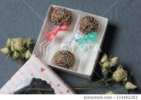 Cake Pops in chocolate with colored sprinkles. Decorated with a ribbon bow. They lie in a gift box, in the lid of which there is a transparent window. Nearby are dried roses. On a gray background. Cake Pops in chocolate with colored sprinkles. Decorated with a ribbon bow. They lie in a gift box, in the lid of which there is a transparent window. Nearby are dried roses. On a gray background. 114402261