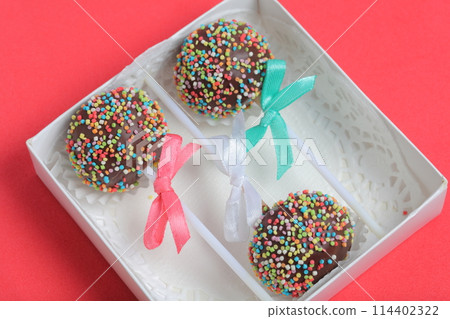Cake Pops in chocolate with colored sprinkles. Decorated with a ribbon bow. They lie in a gift box.  On a red background. 114402322