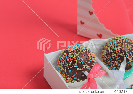 Cake Pops in chocolate with colored sprinkles. Decorated with a ribbon bow. They lie in a gift box, in the lid of which there is a transparent window.  On a red background. 114402323