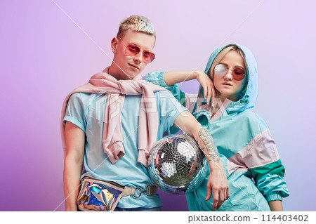 Young fashionable couple of dancers posing with disco ball on violet background 114403402
