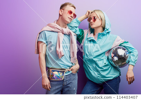 Young fashionable couple of dancers posing with disco ball on violet background Young fashionable couple of dancers posing with disco ball on violet background 114403407