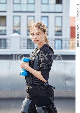 blond woman in Electrical Muscular Stimulation suit standing with blue dumbbell blond woman in Electrical Muscular Stimulation suit standing with blue dumbbell 114404491