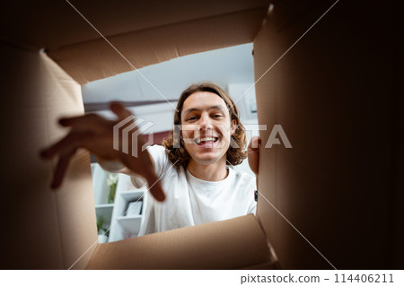 Happy Young Man Reaching Into A Cardboard Box At Home Happy Young Man Reaching Into A Cardboard Box At Home 114406211