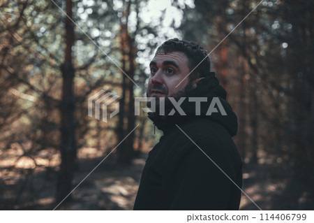 A handsome man looks thoughtfully while standing on a forest path while walking. A man walks through the forest, enjoying the fresh air and thinking. Portrait of a man in the forest. 114406799
