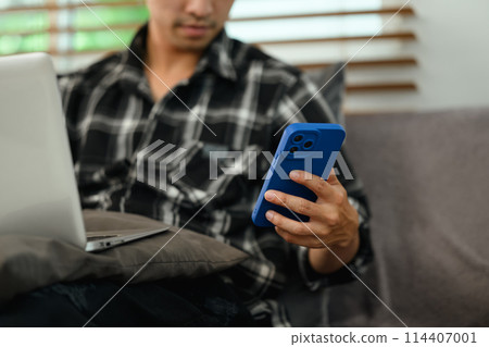 Male freelancer working with laptop and using mobile phone on couch at home 114407001