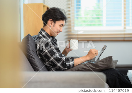 Happy young man drinking coffee and surfing internet on digital tablet at home 114407019