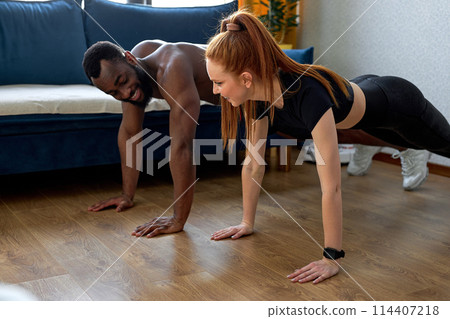 Couple exercising together, redhead woman and black muscular guy ay home workout Couple exercising together, redhead woman and black muscular guy ay home workout 114407218