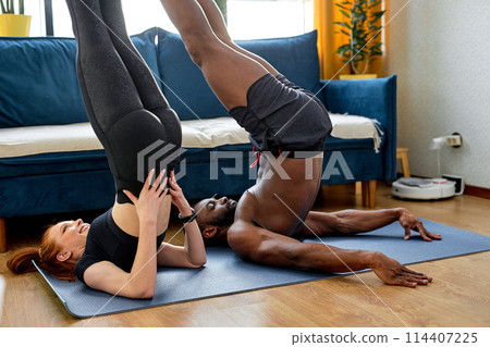 mixed race couple in sportswear are doing exercises at home in a bright cozy living room mixed race couple in sportswear are doing exercises at home in a bright cozy living room 114407225