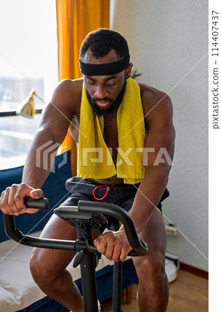 fit muscular man with naked torso riding a stationary bike at home, training workout fit muscular man with naked torso riding a stationary bike at home, training workout 114407437