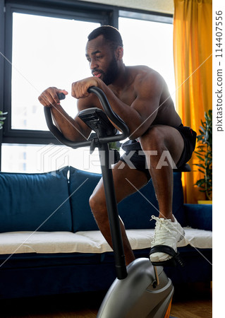 strong black fitness man training on a stationary bike, exercise at home alone strong black fitness man training on a stationary bike, exercise at home alone 114407556