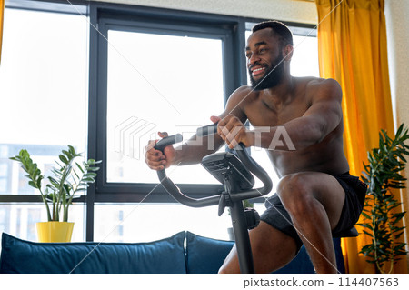Young black fitness man training on a stationary bike, exercise at home alone 114407563