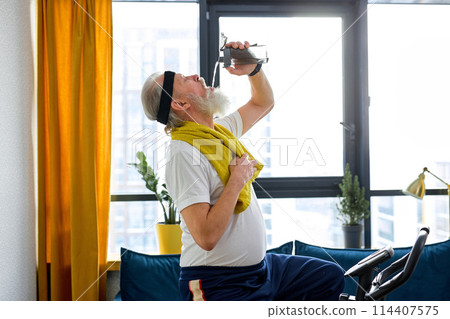 Elderly adult man doing physical cardio exercise on fitness bicycle, drinking water exhausted Elderly adult man doing physical cardio exercise on fitness bicycle, drinking water exhausted 114407575