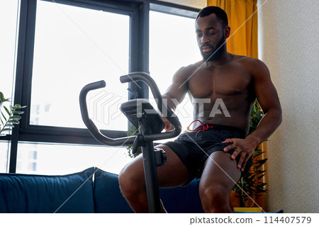 fit muscular man with naked torso riding a stationary bike at home, training workout fit muscular man with naked torso riding a stationary bike at home, training workout 114407579