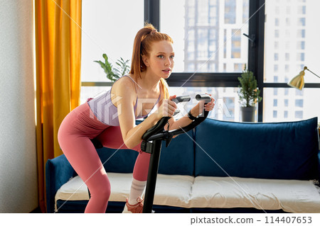 young redhead woman working out on exercise bike or stationary bike at home alone 114407653