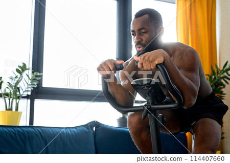 confident muscular man with naked torso riding a stationary bike at home, training workout 114407660