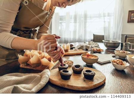 Woman skillfully decorates cupcakes on a wooden board, surrounded by baking supplies and kitchen tools, showcasing the artistry of homemade desserts 114409313