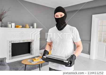 Thief with black balaclava stealing modern expensive laptop from apartment. Masked face. Man burglar stealing tv set from house 114411252
