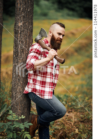 bearded lumberjack worker standing in forest with axe 114411978