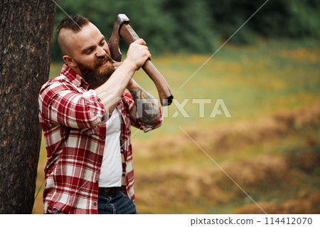 man lumberjack with beard and moustache in red checkered shirt shaves with axe blade on natural background 114412070