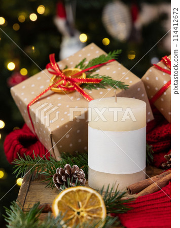 Pillar candle with label near wrapped Christmas gift, red sweater and decor, winter mockup Pillar candle with label near wrapped Christmas gift, red sweater and decor, winter mockup 114412672