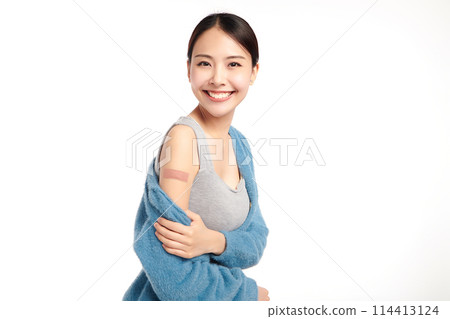 young asian women smiling after getting a vaccine, holding down her shirt sleeve and showing her arm with bandage after receiving vaccination on white background, 114413124