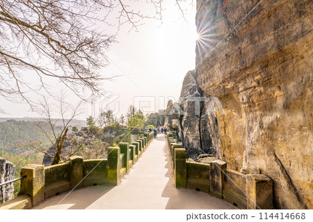 Visitors walk along the historic Bastei Bridge, bathed in sunlight, amidst the rock formations of Saxon Switzerland National Park. Germany 114414668