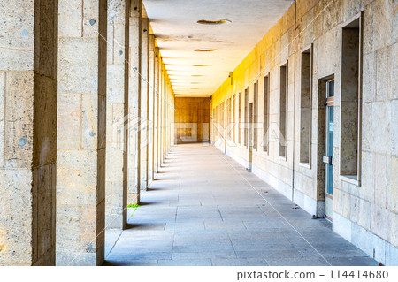 A corridor lined with columns and doors is bathed in sunlight, creating a pattern of light and shadow on the floor. 114414680