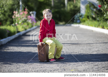 A sad boy sits on an old suitcase on the road. The child is about to leave. A sad boy sits on an old suitcase on the road. The child is about to leave. 114414898