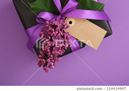 Black craft gift box with empty tag, lilac flowers on violet background. Place for your text Black craft gift box with empty tag, lilac flowers on violet background. Place for your text 114414947