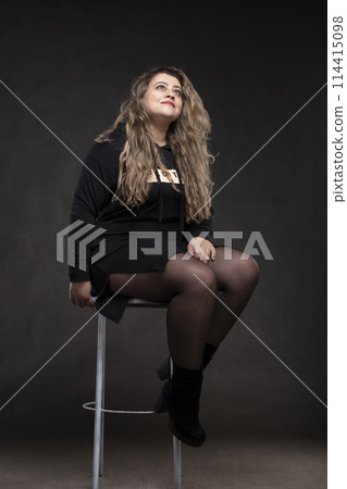 Beautiful fat woman with long curly hair in black clothes on a gray background. Plus size model girl. Beautiful fat woman with long curly hair in black clothes on a gray background. Plus size model girl. 114415098