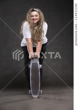 Beautiful fat woman with a skateboard on a gray background looks at the camera and smiles. Beautiful fat woman with a skateboard on a gray background looks at the camera and smiles. 114415100