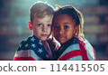 Two small children, a blond boy and a small African-American woman, under the national flag of America. The concept of celebrating Independence Day in America on July 4th 114415505