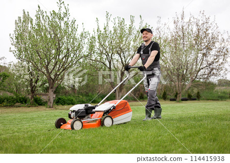 Professional gardener in protective apparel is mowing green grass lawn using modern gasoline cordless lawnmower at backyard. Seasonal landscaping design work. Blooming trees on background 114415938